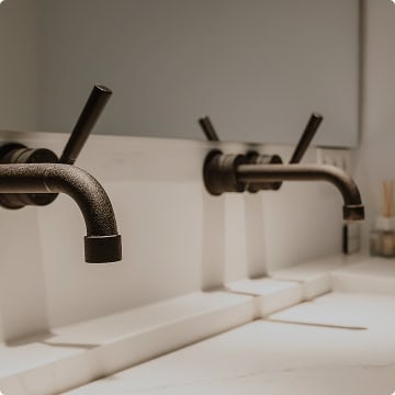 Two modern, wall-mounted black faucets extend over a white sink countertop in a minimalist bathroom setting—an example of the clean craftsmanship Honest Abe Plumbing is known for.