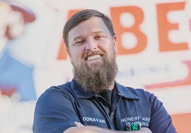 Man with a beard wearing an Honest Abe Plumbing navy blue shirt, standing outdoors and smiling with arms crossed.