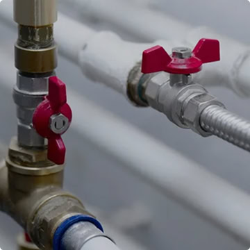 Two metal pipes connected with brass and steel fittings feature red butterfly shutoff valves, with additional white pipes in the background—a quality installation by Honest Abe Plumbing.