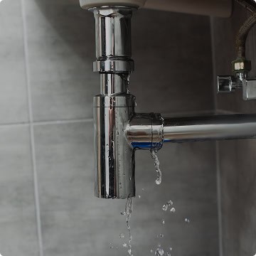 Water is leaking from a shiny metal pipe under a sink against a tiled wall—time to call Honest Abe Plumbing for reliable repairs.