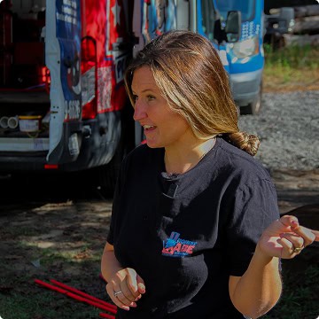 Woman with light brown hair in a black T-shirt stands outdoors near two Honest Abe Plumbing service vans, gesturing with her right hand and looking to the side.