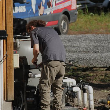 A person in a gray shirt and khaki pants, representing Honest Abe Plumbing, services outdoor equipment near a building, with a work van and gravel in the background.