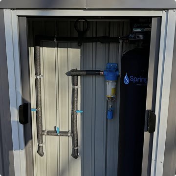 Water filtration system by Honest Abe Plumbing with insulated pipes, control valves, and a blue filter unit installed inside a metal enclosure.